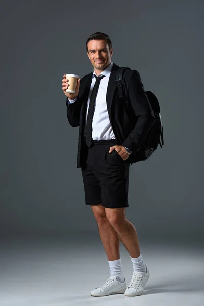 Hombre elegante en pantalones cortos con mochila sosteniendo taza de papel de café sobre fondo gris - foto de stock