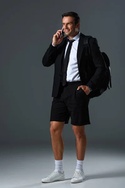 Улыбающийся стильный мужчина в шортах с рюкзаком разговаривая на смартфоне и держа руку в кармане на сером фоне — стоковое фото
