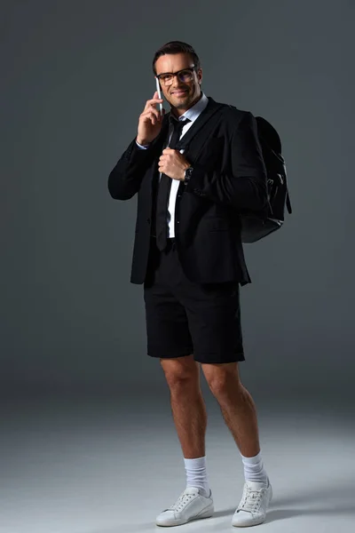 Homem elegante feliz em shorts com mochila falando no smartphone em fundo cinza — Fotografia de Stock