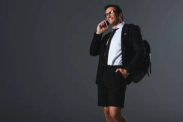Улыбающийся стильный мужчина в шортах с рюкзаком разговаривает по смартфону на сером фоне — стоковое фото