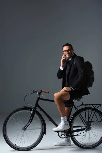 Hombre de moda con mochila sentado en bicicleta hablando en el teléfono inteligente sobre fondo gris - foto de stock