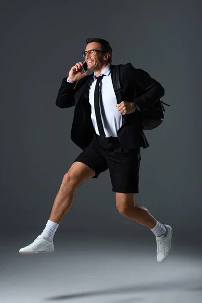Animado homem da moda em óculos falando no smartphone e pulando com mochila no fundo cinza — Fotografia de Stock
