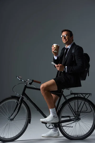 Модный человек с рюкзаком сидит на велосипеде со смартфоном в руке и пьет кофе на сером фоне — стоковое фото