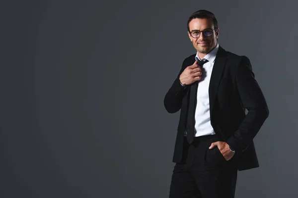 Щасливий бізнесмен в окулярах з рукою в кишені регулюючи краватку ізольовано на сірому фоні — стокове фото