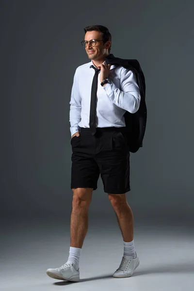 Sonriente hombre elegante con gafas en pantalones cortos sosteniendo chaqueta en el hombro sobre fondo gris - foto de stock