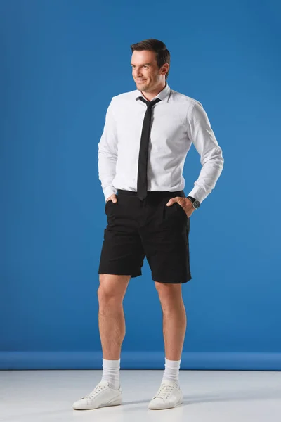 Sonriente hombre elegante en pantalones cortos con las manos en los bolsillos sobre fondo azul - foto de stock