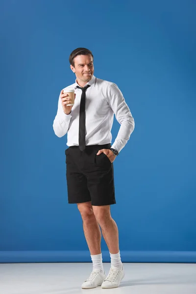 Schöner lächelnder Mann mit Einweg-Kaffeetasse, während er mit der Hand in der Hosentasche auf blauem Grund steht — Stockfoto