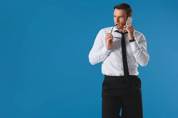 Homem sério mordendo óculos enquanto conversa por smartphone e olhando para longe isolado no azul — Fotografia de Stock