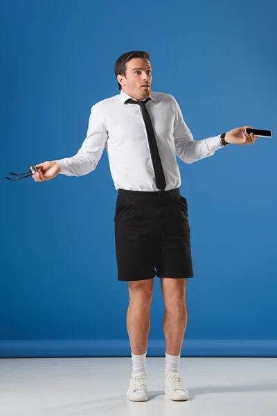 Shocked man holding smartphone and eyeglasses and looking away on blue — Stock Photo