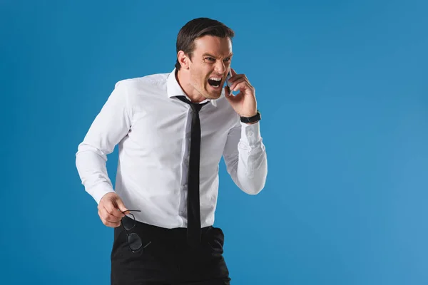 Сердитий чоловік тримає окуляри і кричить під час розмови зі смартфоном ізольовано на синьому — стокове фото