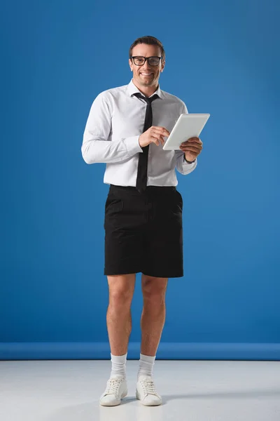 Guapo fetichista en gafas y pantalones cortos usando tableta digital y sonriendo a la cámara en azul — Stock Photo