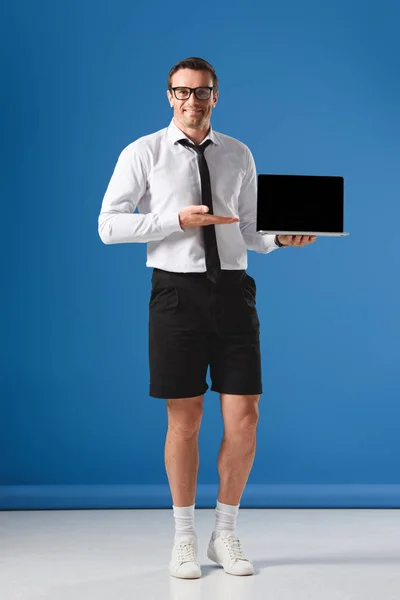 Guapo hombre de negocios en pantalones cortos mostrando portátil con pantalla en blanco y sonriendo a la cámara en azul - foto de stock