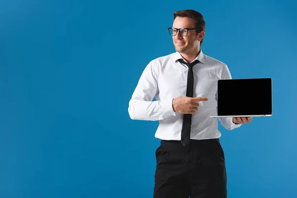 Hombre sonriente en gafas apuntando con el dedo a la computadora portátil con la pantalla en blanco y mirando hacia otro lado aislado en azul - foto de stock
