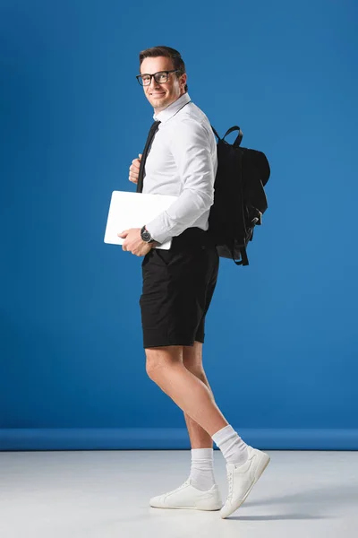 Buisnessman feliz en gafas y pantalones cortos que sostienen el ordenador portátil y sonriendo a la cámara en azul - foto de stock