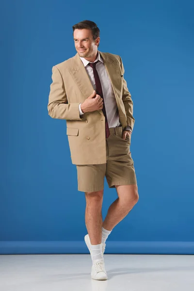 Vista de longitud completa de hombre sonriente guapo de pie con la mano en el bolsillo y mirando hacia otro lado en azul - foto de stock
