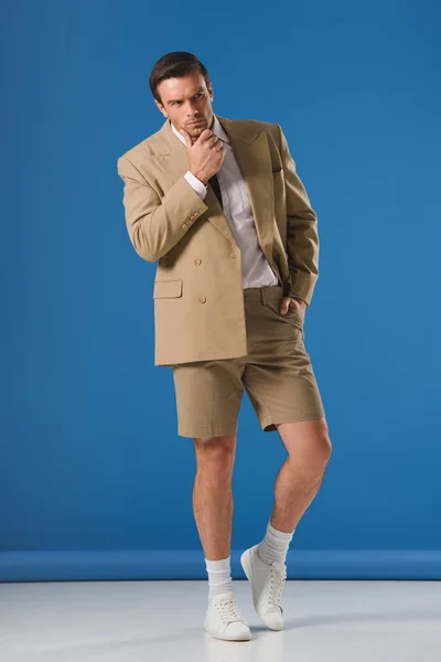 Thoughtful man in shorts standing with hand on chin and looking away on blue — Stock Photo