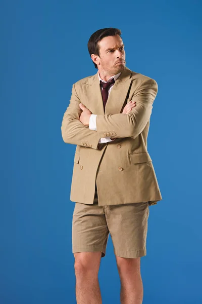Hombre molesto en pantalones cortos de pie con los brazos cruzados y mirando hacia otro lado aislado en azul - foto de stock
