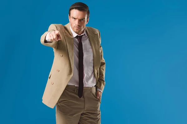 Serious man standing with hand in pocket and pointing at camera isolated on blue — Stock Photo