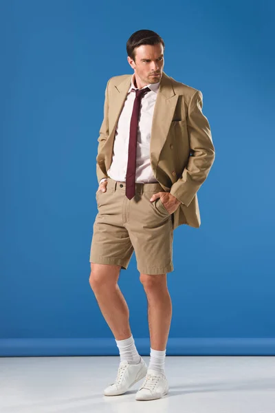 Full length view of handsome man in suit jacket and shorts standing with hands in pockets and looking away on blue — Stock Photo