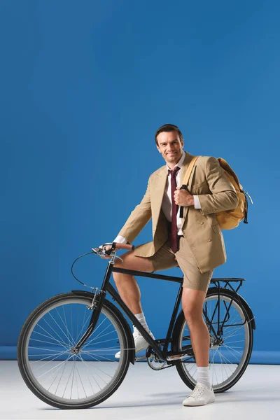 Hombre guapo con mochila sentado en bicicleta y sonriendo a la cámara en azul - foto de stock