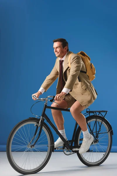 Усміхнений чоловік у піджак і шорти їзда на велосипеді і фотографіях хтось дивитися вбік на блакитному — стокове фото