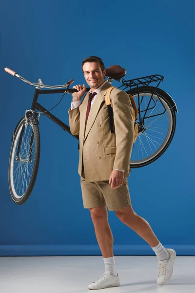 Vista completa de hombre guapo llevando bicicleta y sonriendo a la cámara en azul - foto de stock
