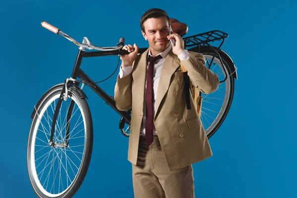 Sonriente hombre llevando bicicleta y hablando por teléfono inteligente aislado en azul - foto de stock