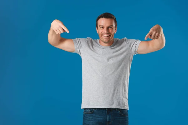 Hombre feliz señalando con los dedos y la camiseta y sonriendo a la cámara aislado en azul - foto de stock