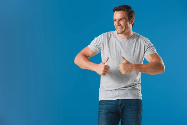 Hombre sonriente guapo mostrando los pulgares hacia arriba y mirando hacia otro lado aislado en azul - foto de stock