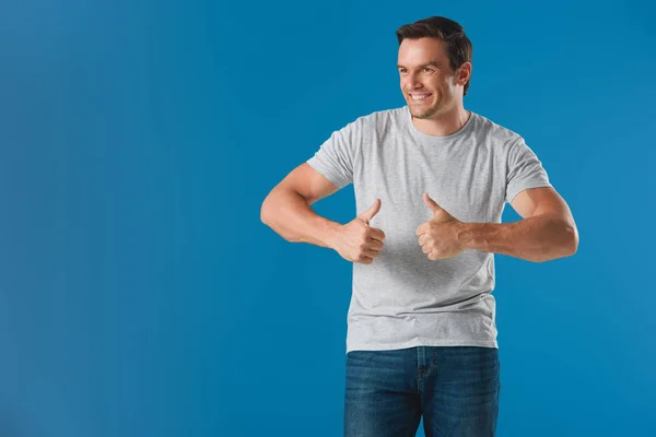 Bonito sorrindo homem mostrando polegares para cima e olhando para longe isolado no azul — Fotografia de Stock