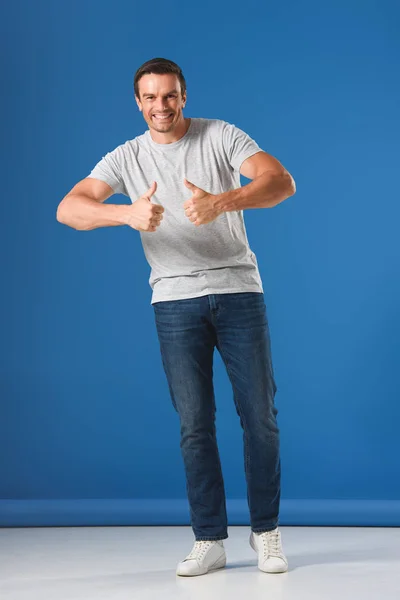 Feliz hombre guapo mostrando pulgares hacia arriba y sonriendo a la cámara en azul - foto de stock