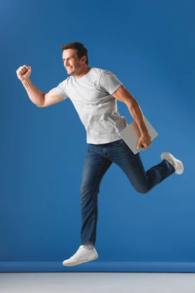 Fröhlicher Mann hält Laptop in der Hand und springt auf blau — Stockfoto