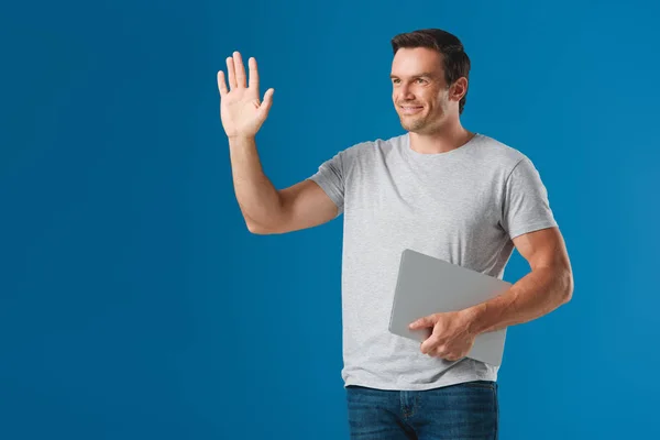 Щасливий чоловік тримає ноутбук і махає рукою, дивлячись ізольовано на синьому — стокове фото