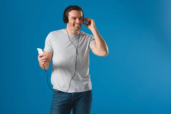 Homme heureux dans les écouteurs écouter de la musique avec smartphone et regarder loin isolé sur bleu — Photo de stock