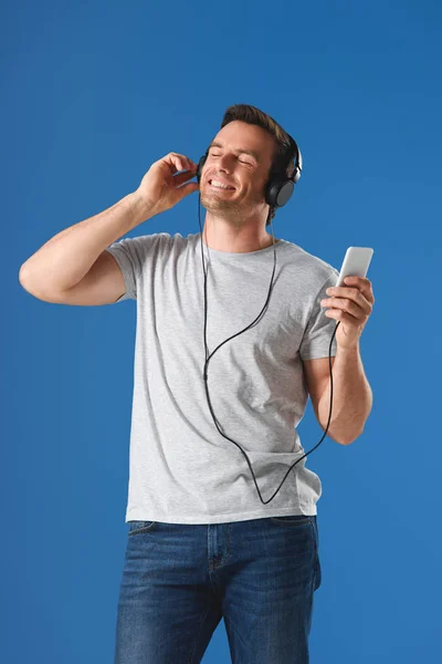 Homme souriant dans les écouteurs écouter de la musique avec les yeux fermés et tenant smartphone isolé sur bleu — Photo de stock