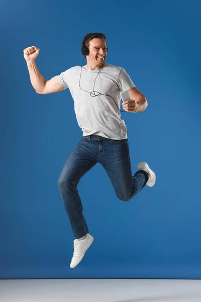 Fröhlicher Mann mit Kopfhörer hält Smartphone in der Hand und springt auf blau — Stockfoto