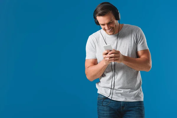 Uomo sorridente in cuffia utilizzando smartphone isolato su blu — Foto stock