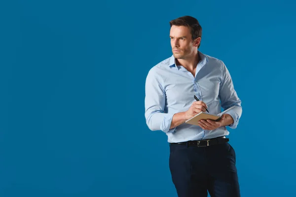 Handsome businessman taking notes in notepad and looking away isolated on blue — Stock Photo