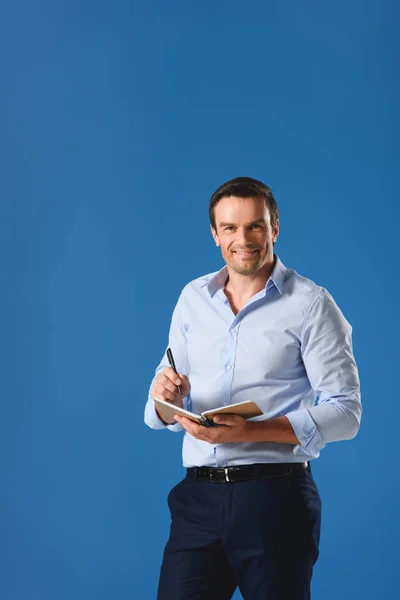 Guapo hombre de negocios escribiendo en cuaderno y sonriendo a la cámara aislado en azul - foto de stock