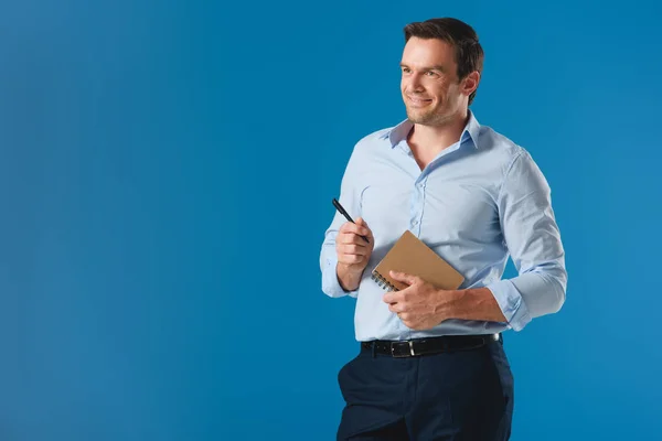 Apuesto hombre de negocios sonriente sosteniendo el cuaderno y la pluma, mirando hacia otro lado aislado en azul - foto de stock