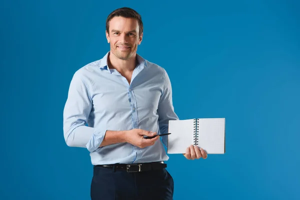 Schöner Geschäftsmann zeigt auf leeres Notizbuch und lächelt in die Kamera isoliert auf blauem Grund — Stockfoto