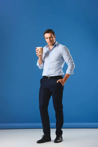 Handsome man holding disposable coffee cup and looking at camera on blue — Stock Photo