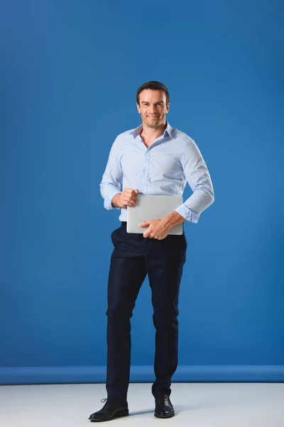 Full length view of handsome man holding laptop and smiling at camera on blue — Stock Photo