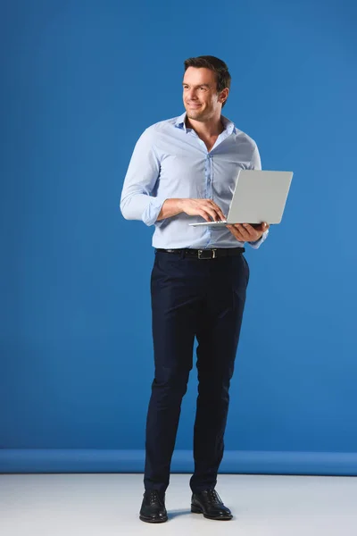 Вид в полный рост на красивого улыбающегося человека, держащего ноутбук и отводящего взгляд в сторону на голубом фоне — стоковое фото