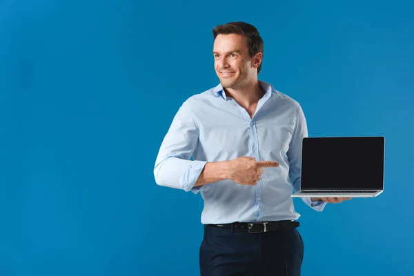 Bel homme souriant pointant du doigt à l'ordinateur portable avec écran blanc et regardant loin isolé sur bleu — Photo de stock