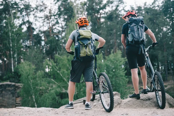 Vista trasera de ciclistas extremos masculinos en cascos protectores de pie con bicicletas de montaña en el bosque - foto de stock