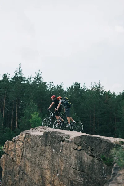 Vista lejana de ciclistas extremos masculinos en cascos protectores montados en bicicletas de montaña sobre un acantilado rocoso en el bosque - foto de stock