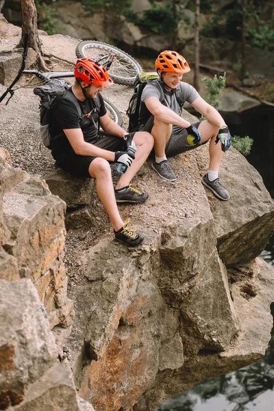 Amigos sonrientes ciclistas con mochilas descansando con una botella deportiva de agua y manzana en un acantilado rocoso cerca del río en el bosque - foto de stock