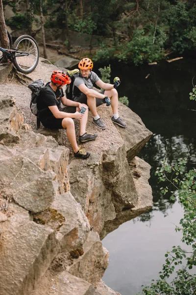 Vista ad alto angolo di amici ciclisti con zaini a riposo con bottiglia d'acqua sportiva e mela sulla scogliera rocciosa vicino al fiume nella foresta — Foto stock