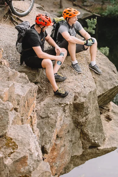 Cyclistes masculins souriants reposant avec une bouteille d'eau sportive et de pomme sur une falaise rocheuse — Photo de stock